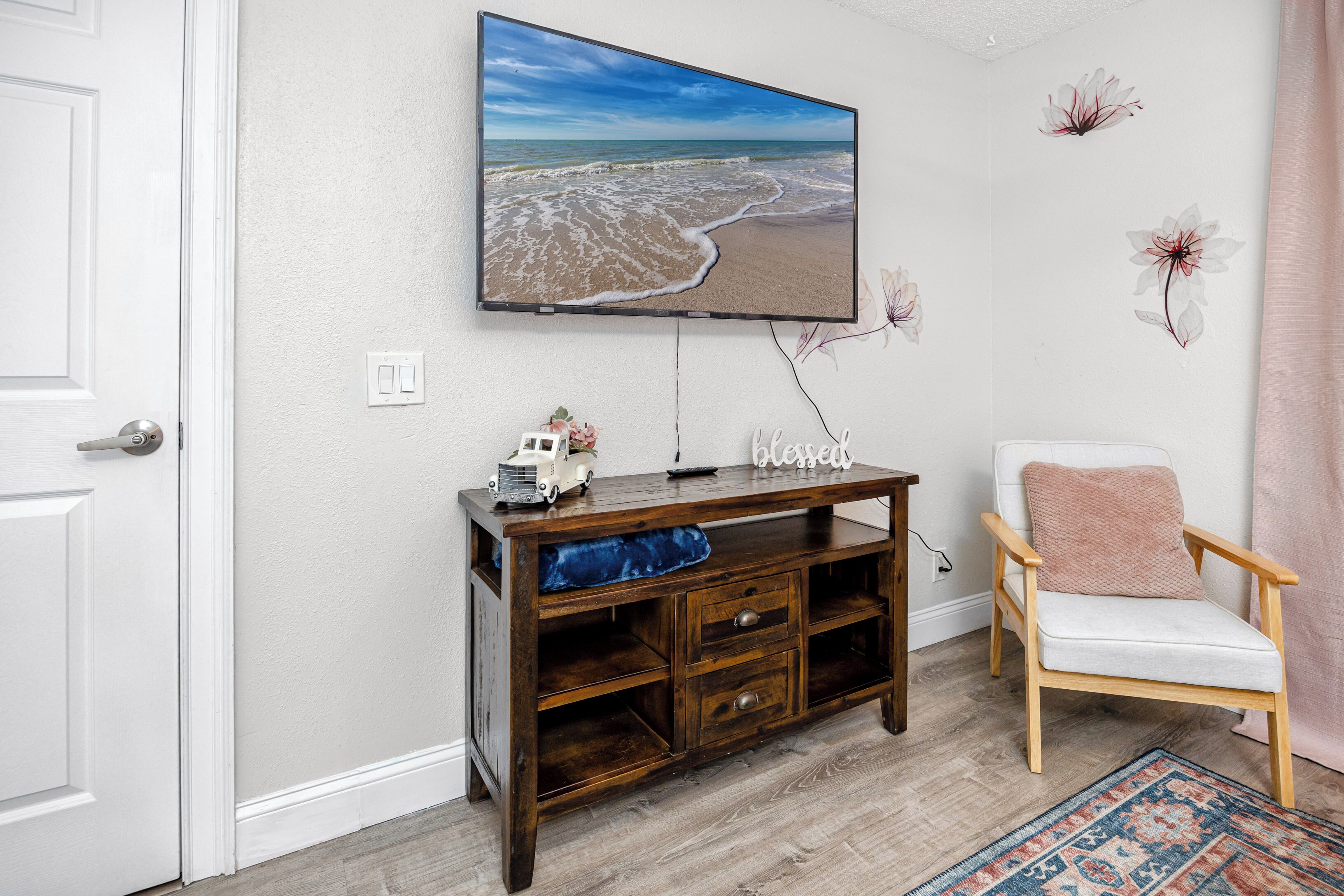 A living room with a television mounted on the wall, a wooden entertainment unit, a white armchair, and a patterned rug.