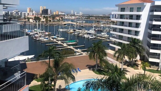 A marina with boats docked, a pool area with palm trees, and modern buildings in the background. - Depas Veleros (Mazatlán)