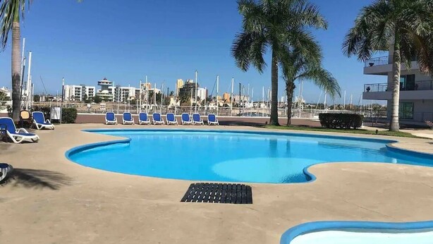 Piscina con sillas de descanso y palmeras, rodeada de edificios y un cielo despejado.