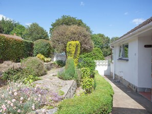 A garden with a stone pathway, various plants, and a white building with a window and a door.