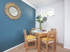 A dining area with a blue wall, a sunburst mirror, wooden chairs with yellow cushions, a table with a white placemat, and a vase with flowers. - 2 bedroom accommodation in Brixham (Brixham)