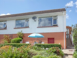 A two-story building with a satellite dish, a red door, and a small garden area with a blue umbrella. - 2 bedroom accommodation in Brixham (Brixham)