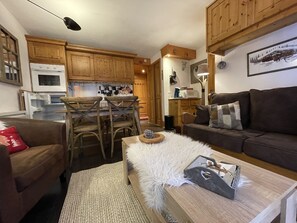 Una sala de estar acogedora con un sofá marrón, un taburete blanco y mullido, y una mesa de centro de madera. Hay una zona de comedor con sillas y una mesa de madera, y una cocina con armarios de madera y un microondas.