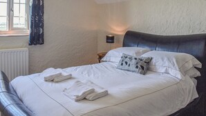 Una cama bien tendida con sábanas blancas y toallas dobladas, cabecera oscura y una lámpara de noche.