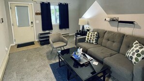 Ein Wohnzimmer mit Sofa, Sessel, Couchtisch und Lampe. Eine Tür und ein Fenster mit Vorhängen sind ebenfalls vorhanden.