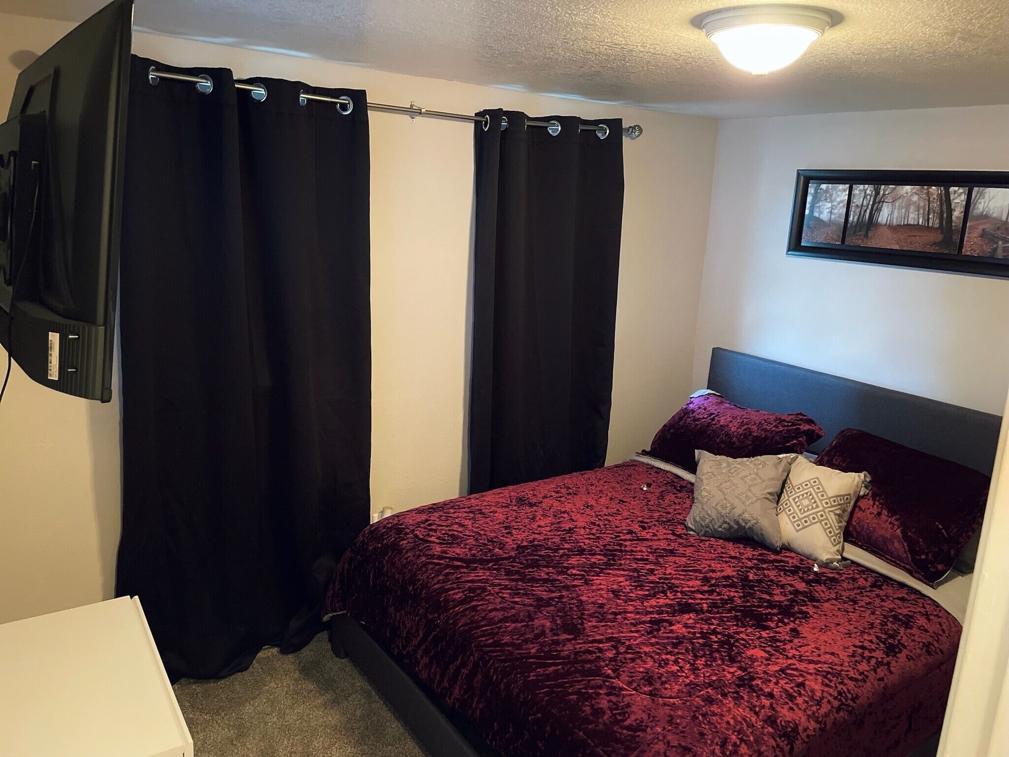A bedroom with a bed, black curtains, a mounted TV, and a framed picture on the wall.