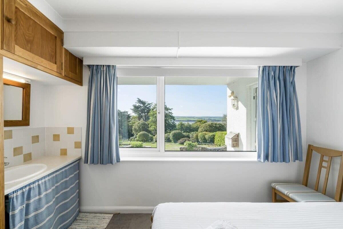 Een slaapkamer met een bed, een raam met gordijnen, een houten hoofdeinde en uitzicht op groen.