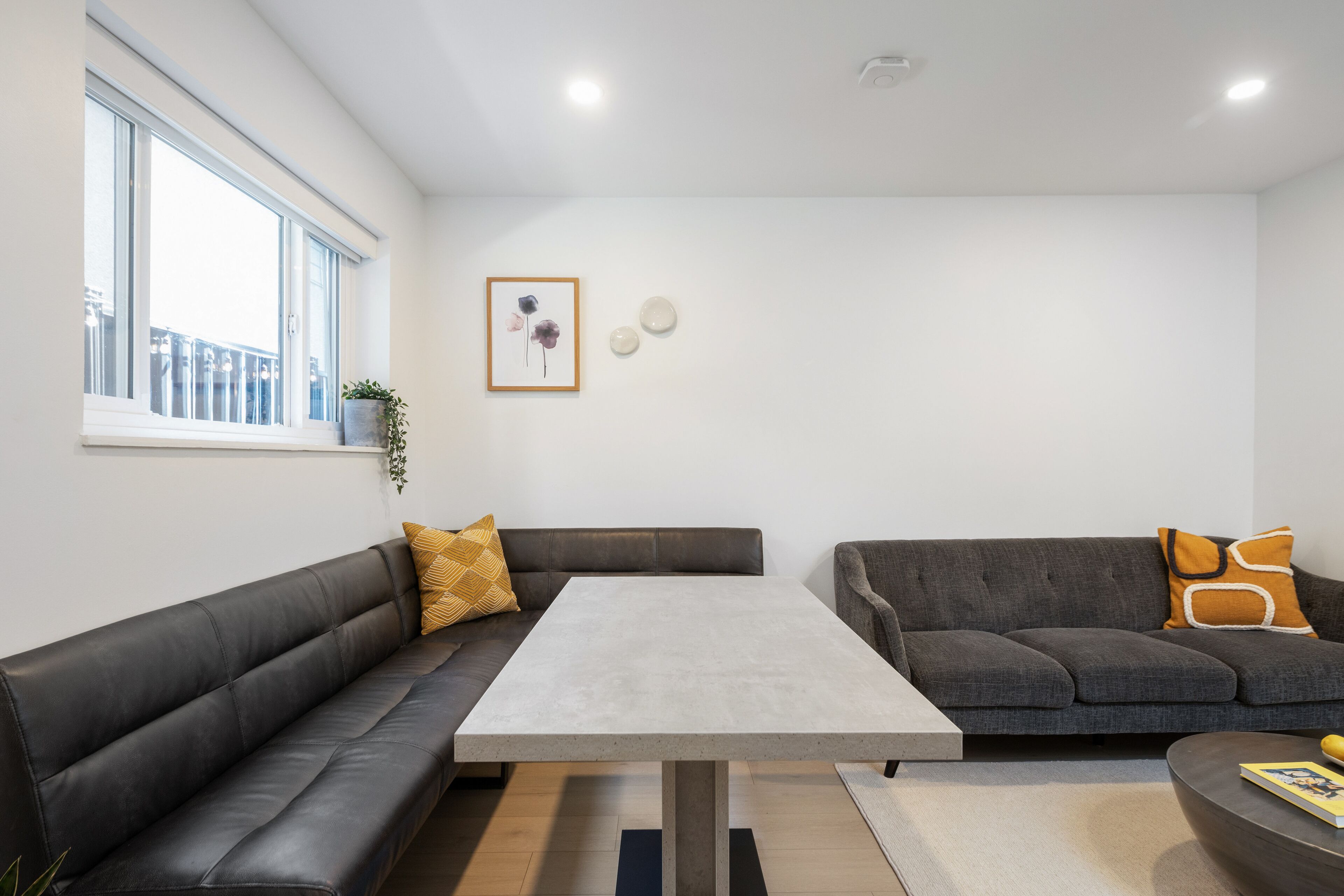 A modern living room with a grey sofa, a matching armchair, a coffee table, and a dining table. There is a window with a view of the outside, a framed picture on the wall, and a small plant on the window sill.