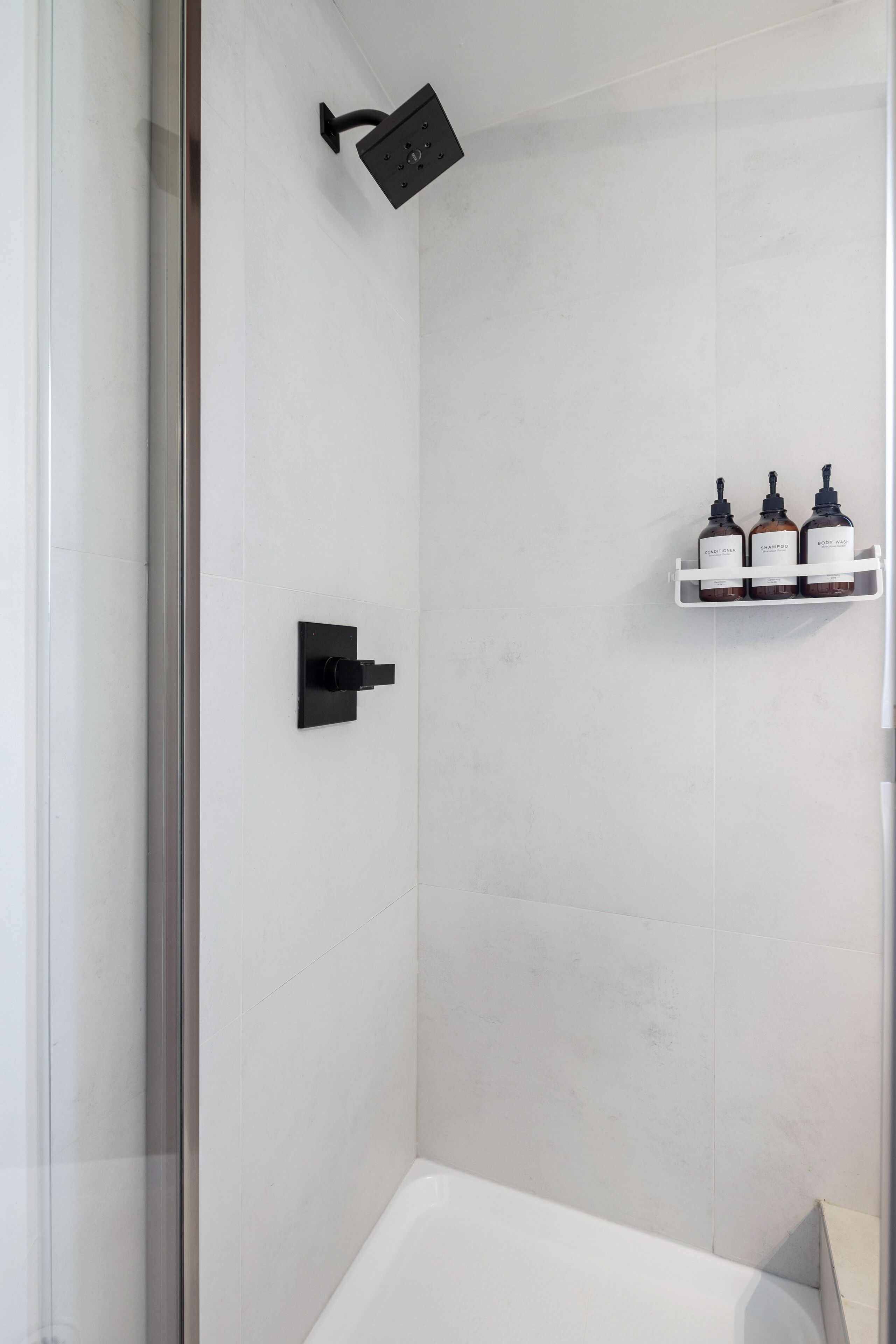 Ein modernes Badezimmer mit einer Duschkabine aus Glas, einer weißen Badewanne und einem Ablagefach mit drei Shampoo-Flaschen.