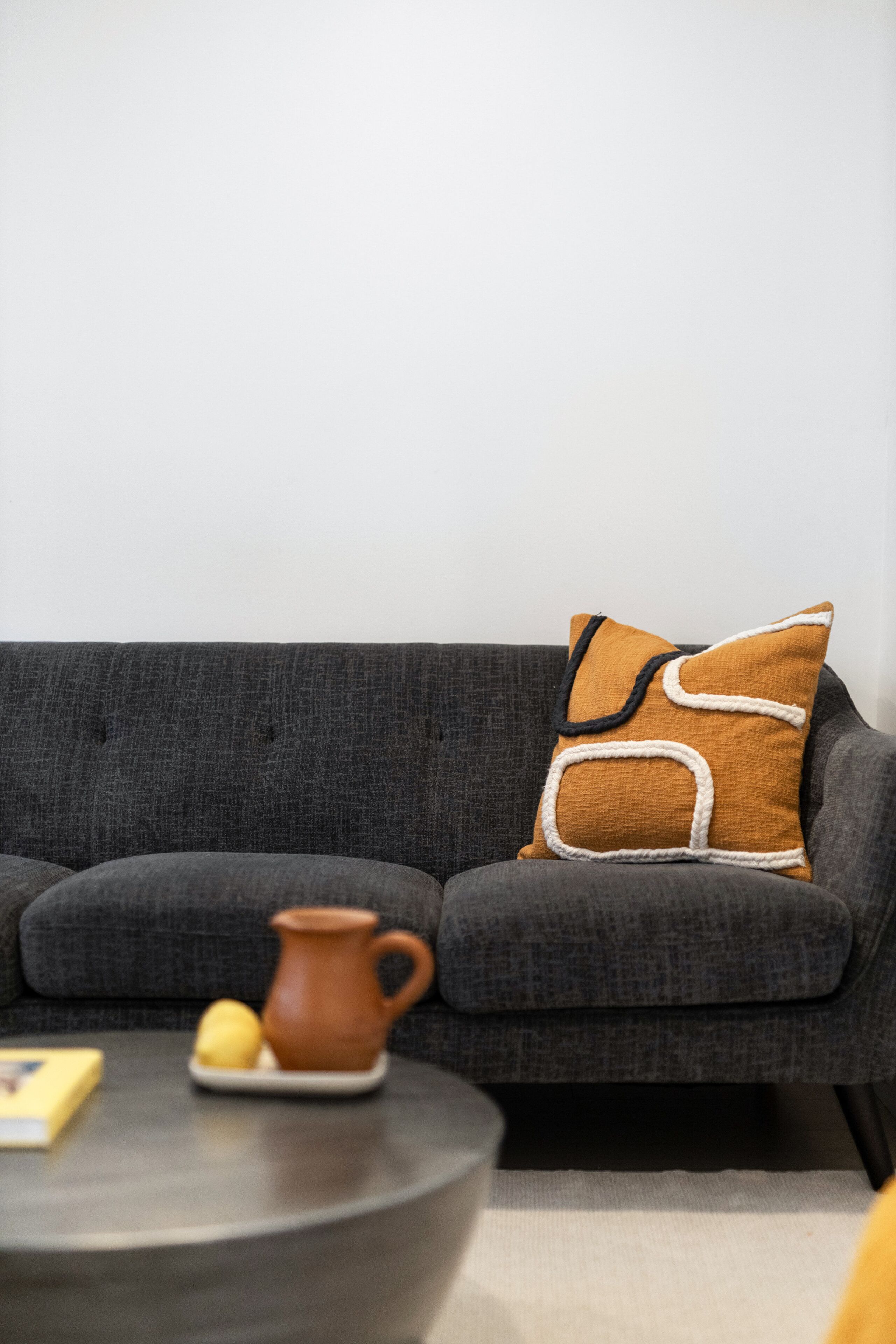 A grey sofa with a brown and white pillow, a small table with a jug and lemons, and a book.