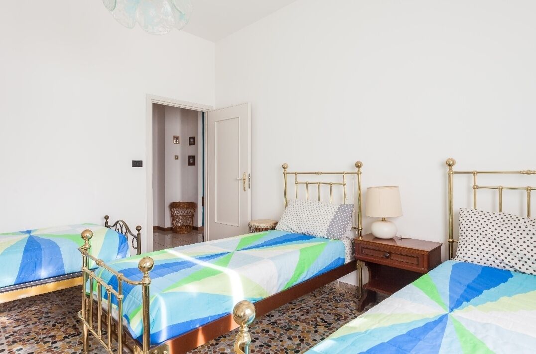 A bedroom with two beds, each with a patterned bedspread, a bedside table with a lamp, and a door leading to another room.