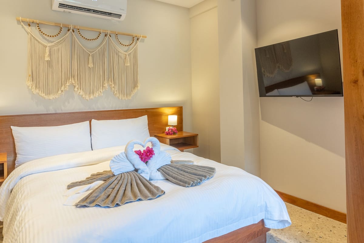 A neatly made bed with a heart-shaped towel arrangement, a wooden headboard, a bedside table with a lamp, and a TV mounted on the wall.