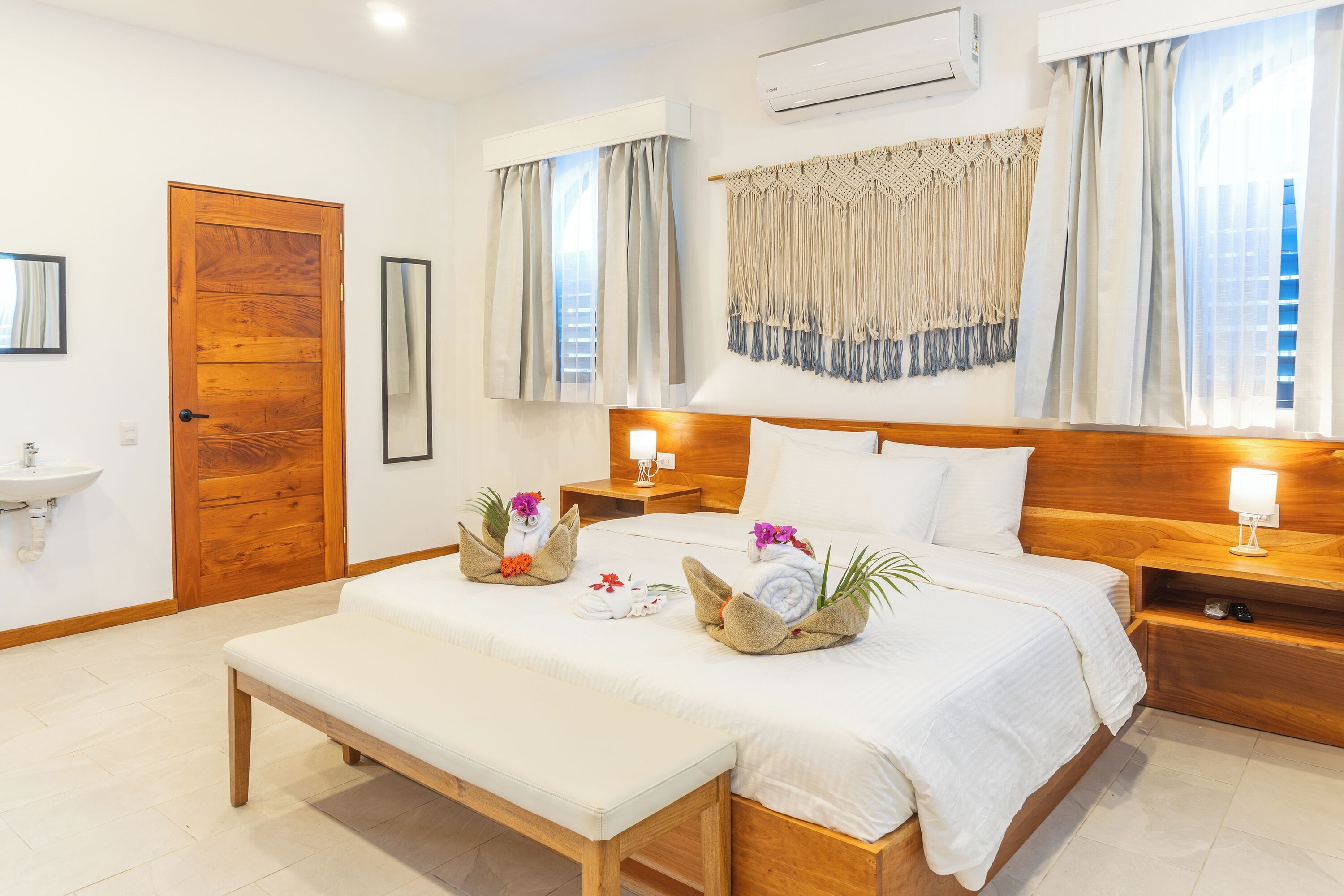 Habitación de hotel con cama de madera, un banco, una mesita de noche con lámpara, aire acondicionado de pared y una ventana con cortinas.