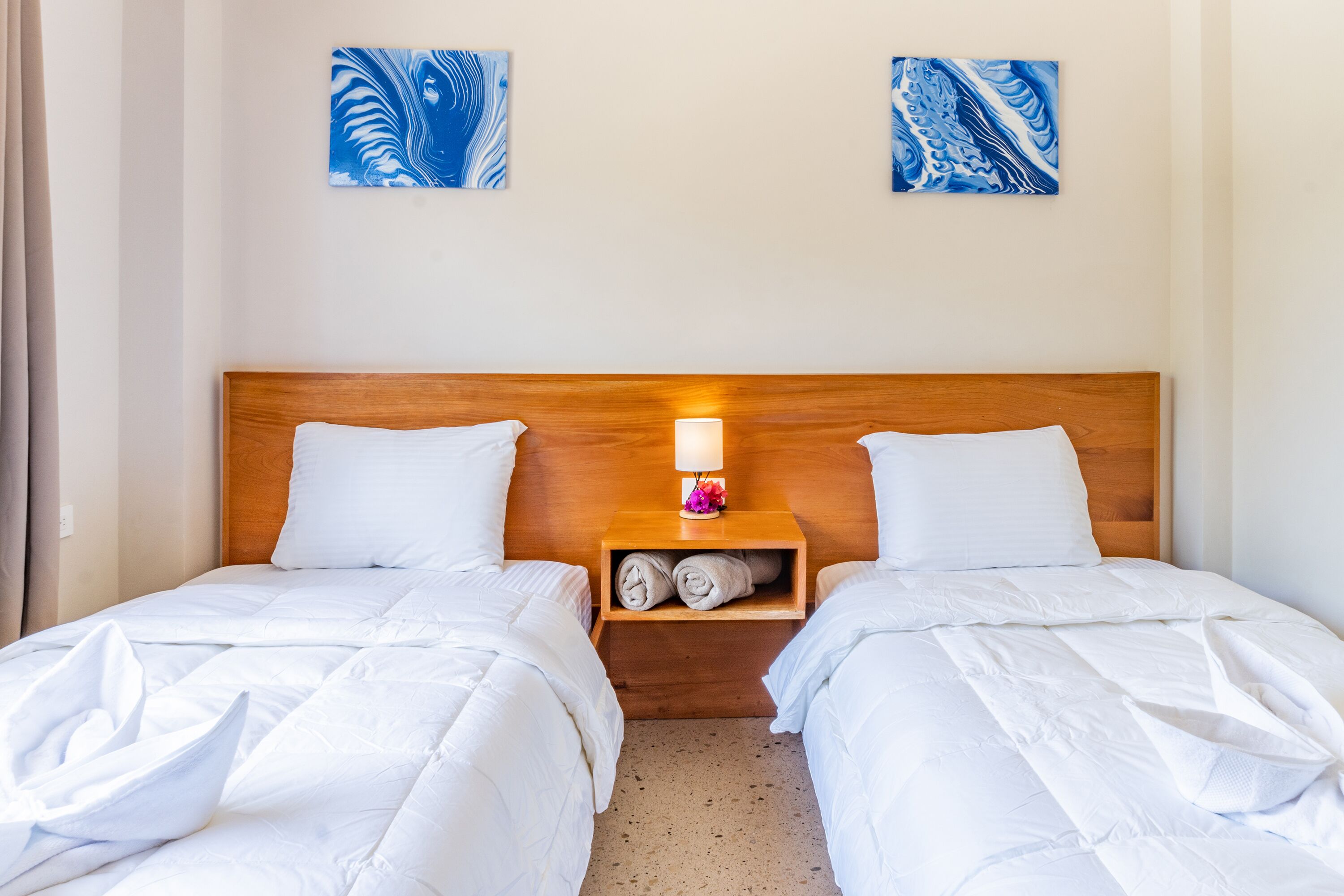 Una cama doble con ropa de cama blanca y cabecera de madera. Hay una mesita de noche con una lámpara y una toalla doblada. Dos cuadros abstractos azules están en la pared sobre la cama.