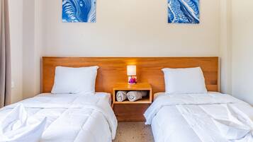 Una cama doble con ropa de cama blanca y cabecera de madera. Hay una mesita de noche con una lámpara y una toalla doblada. Dos cuadros abstractos azules están en la pared sobre la cama.