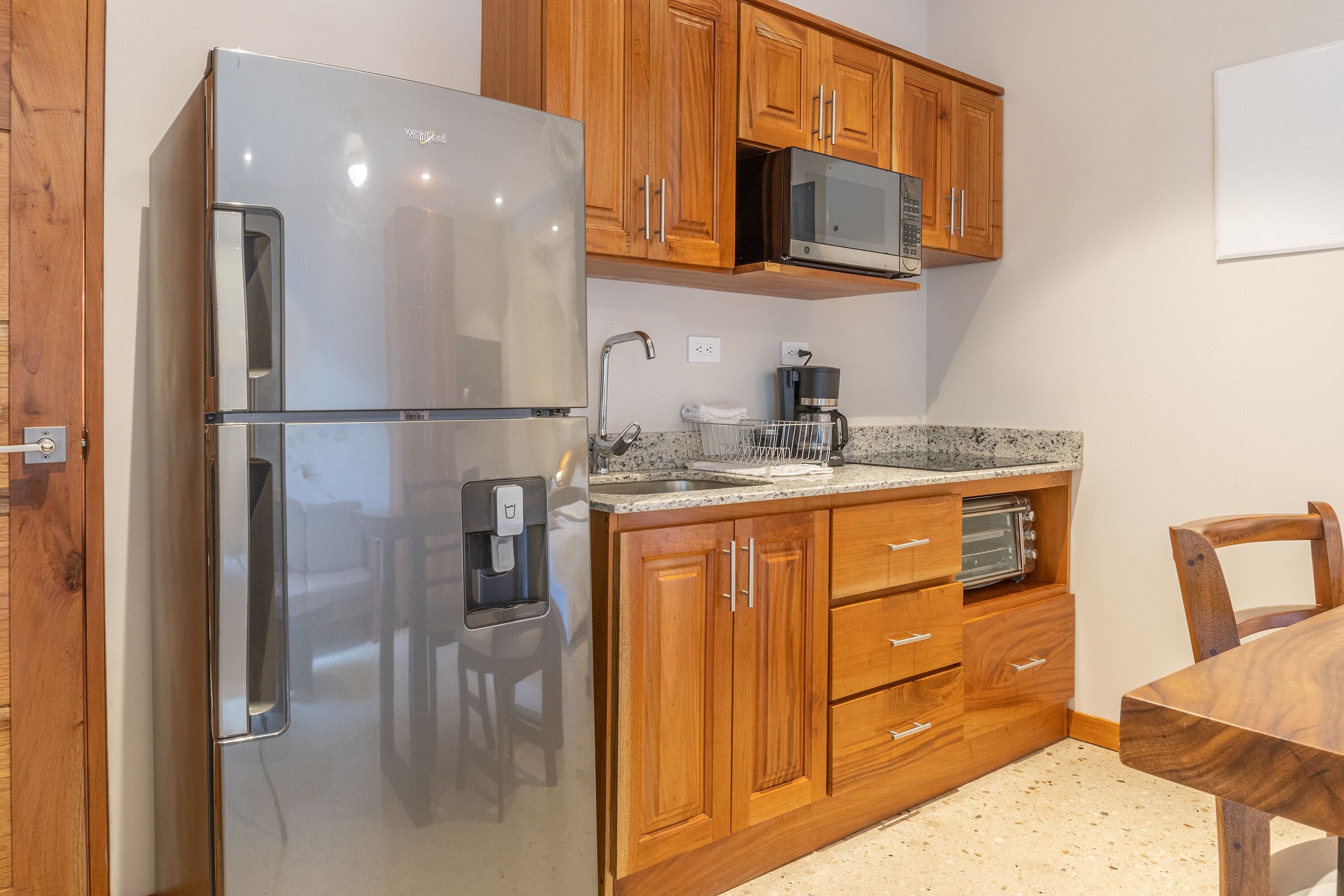 A compact kitchen with wooden cabinets, a stainless steel refrigerator, a microwave, a coffee maker, and a toaster.