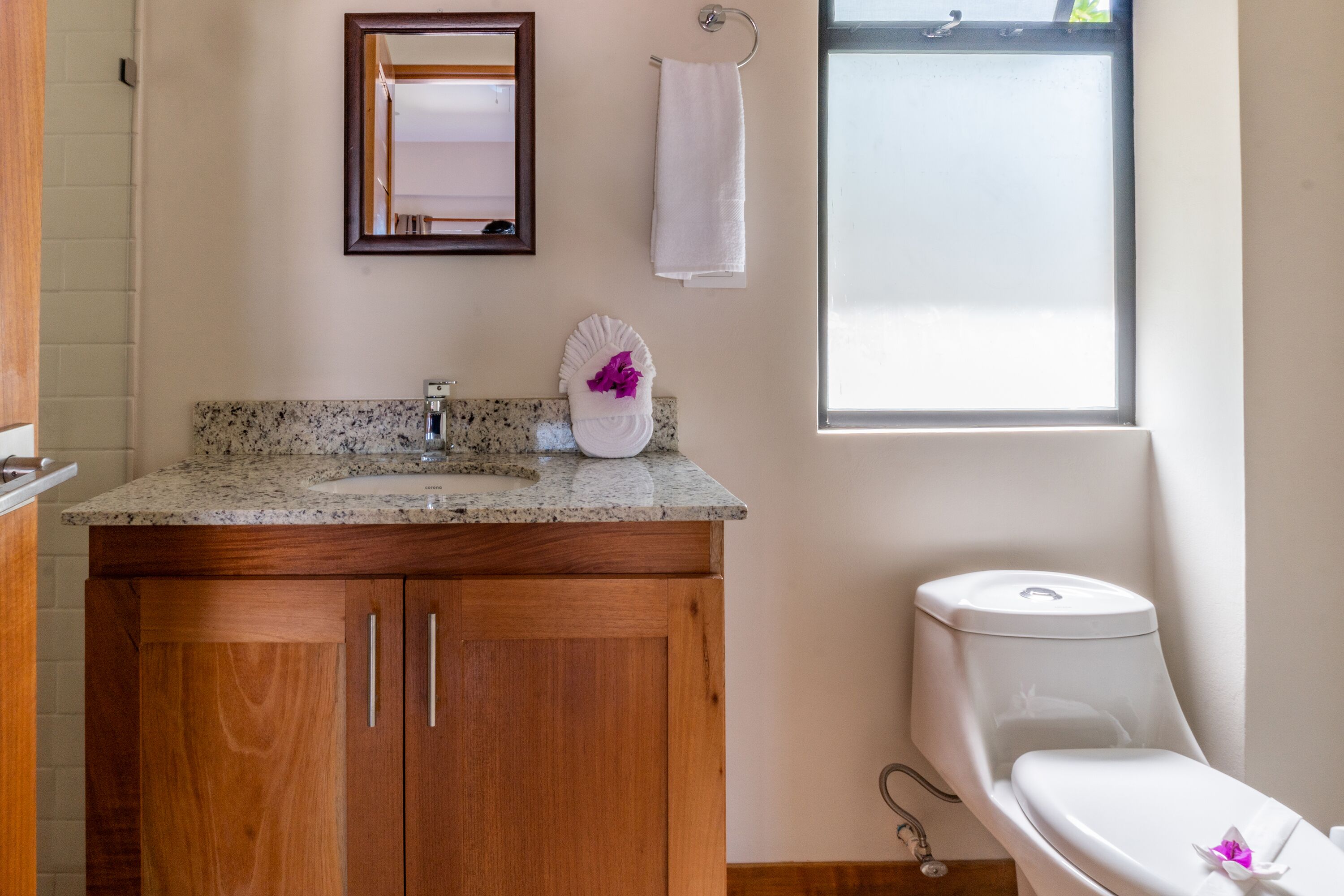 Un baño con encimera de granito, armarios de madera, inodoro blanco, espejo y ventana.