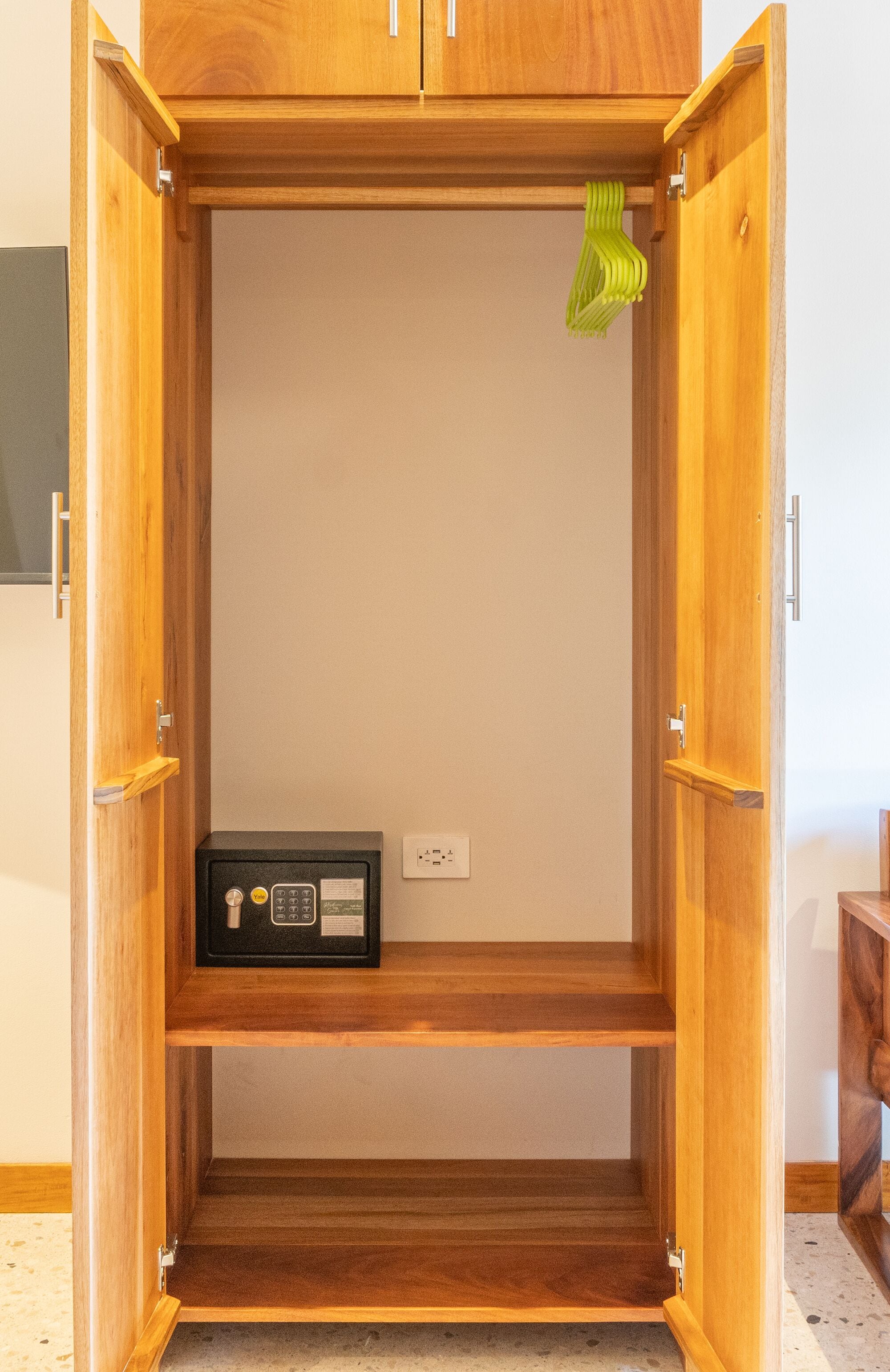 A wooden wardrobe with two shelves and a hanging rod, containing a black safe and a green hanger.