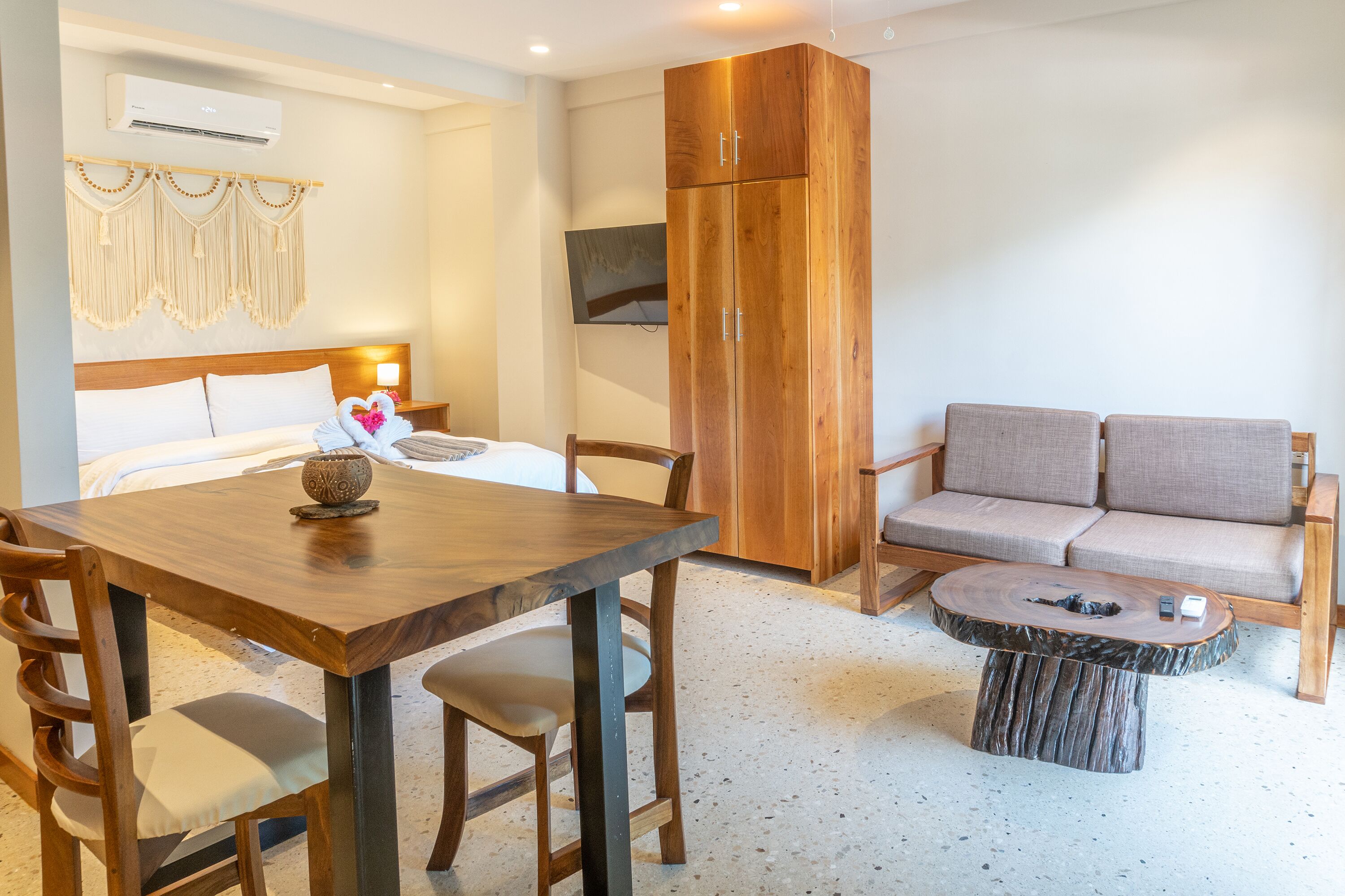A hotel room with a wooden dining table, a sofa, a TV mounted on the wall, and a wardrobe.