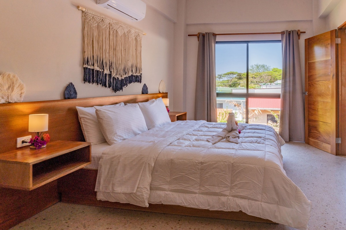 A neatly made bed with white linens, a wooden headboard, and a bedside table with a lamp and flowers.