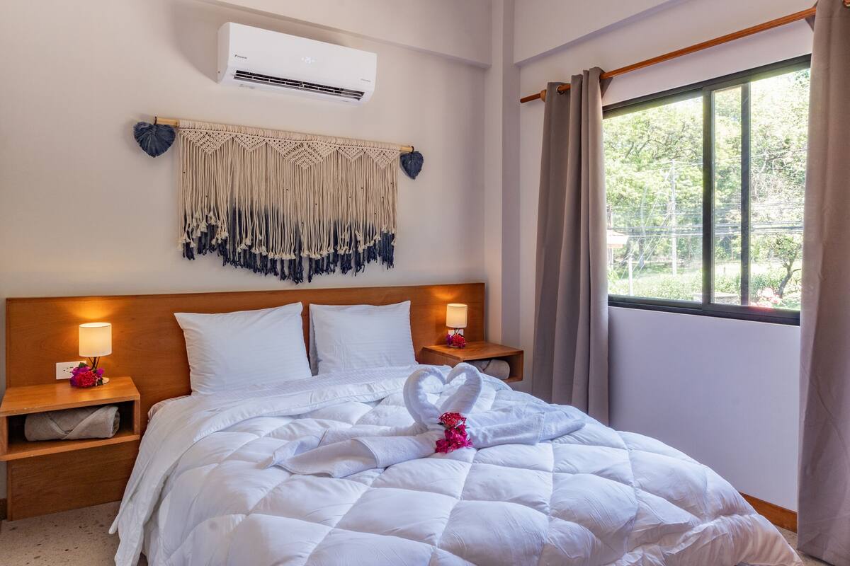 A neatly made bed with white bedding and a heart-shaped towel arrangement, wooden headboard, bedside lamps, a macrame wall hanging, and a window with curtains.