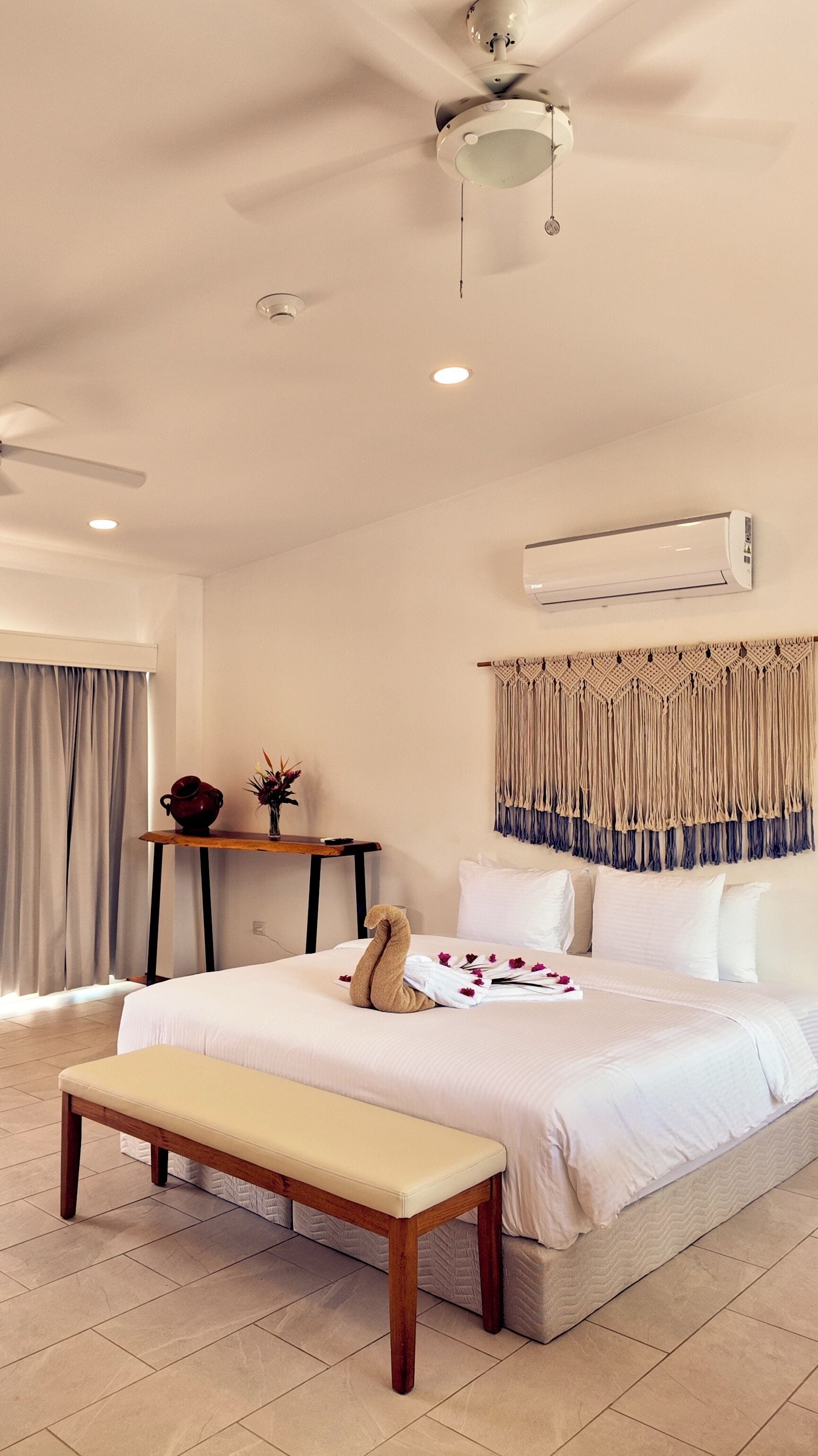 A hotel room with a bed, a bench, a ceiling fan, an air conditioning unit, and a decorative macramé wall hanging.