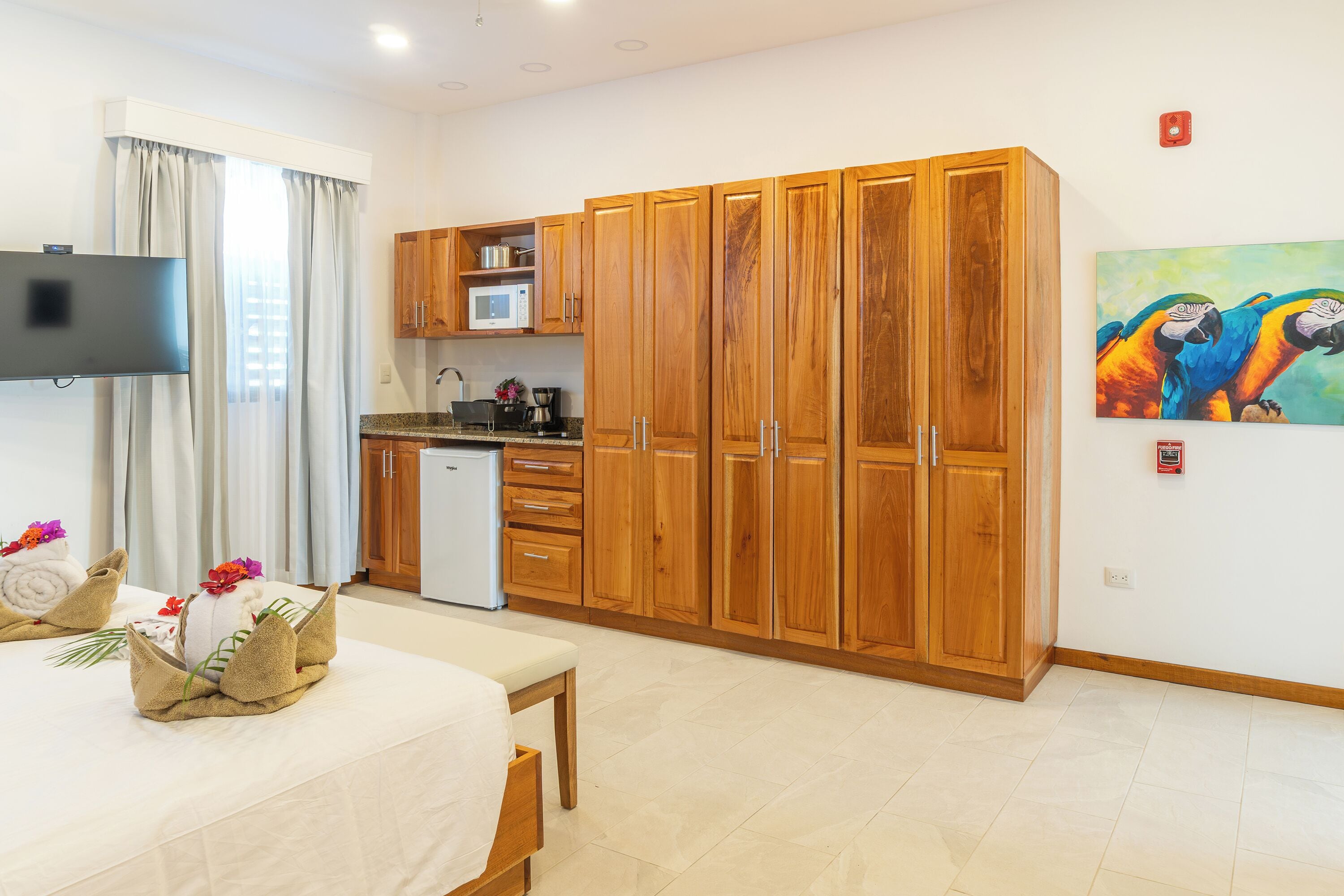 Habitación de hotel con cama, una kitchenette pequeña, armarios de madera, televisor y un cuadro de loros.