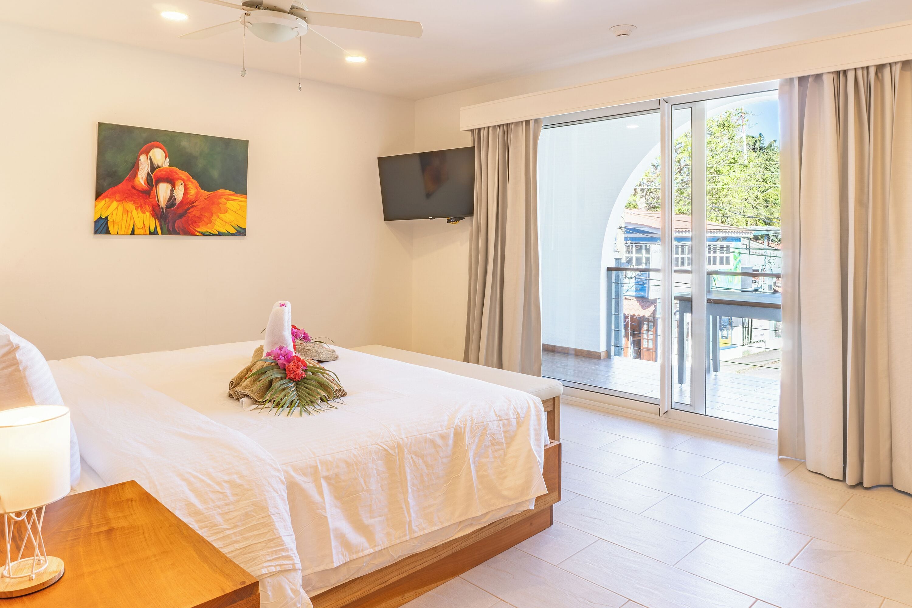 Habitación de hotel con cama, televisor de pantalla plana, balcón con cortinas y un cuadro de loros.
