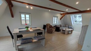 A spacious living area with a wooden dining table, a sofa, and a chair. There are two windows providing natural light. - Vacation apartment in a renovated monument (max. 5 persons) (Bischofswerda)