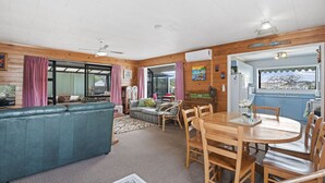 A cozy living room with a blue sofa, a dining table with chairs, and a kitchen area visible through an open doorway. - Beachglen - Snells Beach Holiday Home (Snells Beach)
