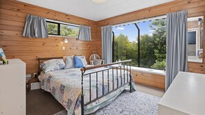 A bedroom with a wooden ceiling, a bed with a metal frame, a bedside table, and a window with curtains. - Beachglen - Snells Beach Holiday Home (Snells Beach)