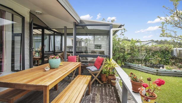 A wooden deck with a table and chairs, surrounded by potted plants and a garden. - Beachglen - Snells Beach Holiday Home (Snells Beach)
