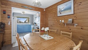 A wooden interior with a round dining table, chairs, a kitchen area, and a framed picture on the wall. - Beachglen - Snells Beach Holiday Home (Snells Beach)