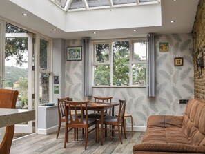 A dining area with a table and chairs, a sofa, and a view of greenery through large windows. - 3 bedroom accommodation in Chesterfield (Chesterfield)