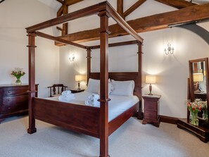 A four-poster bed with a wooden frame, a nightstand with a lamp, a dresser, and a mirror with a vase of flowers.