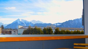 Un balcón con vistas a montañas nevadas, una banca de madera y un edificio con techo plano al frente.