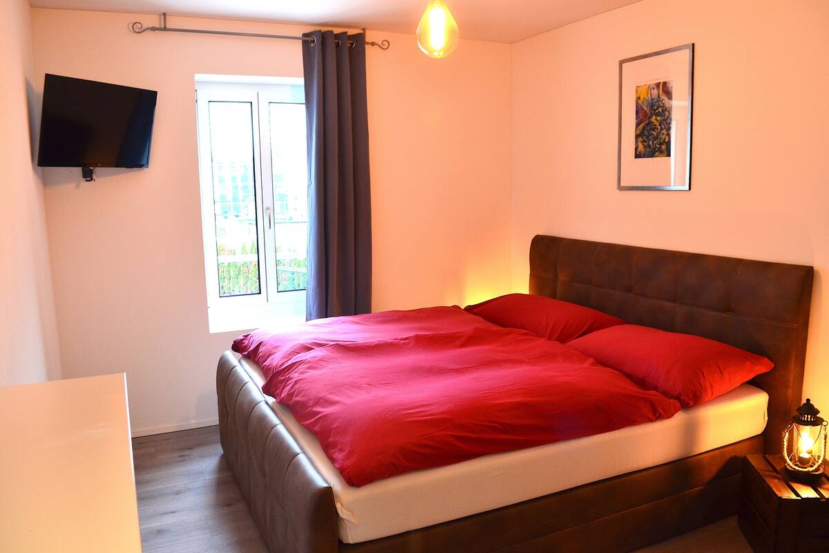 A bedroom with a bed, a flat-screen TV, a window with curtains, a framed picture, and a bedside table with a lantern.
