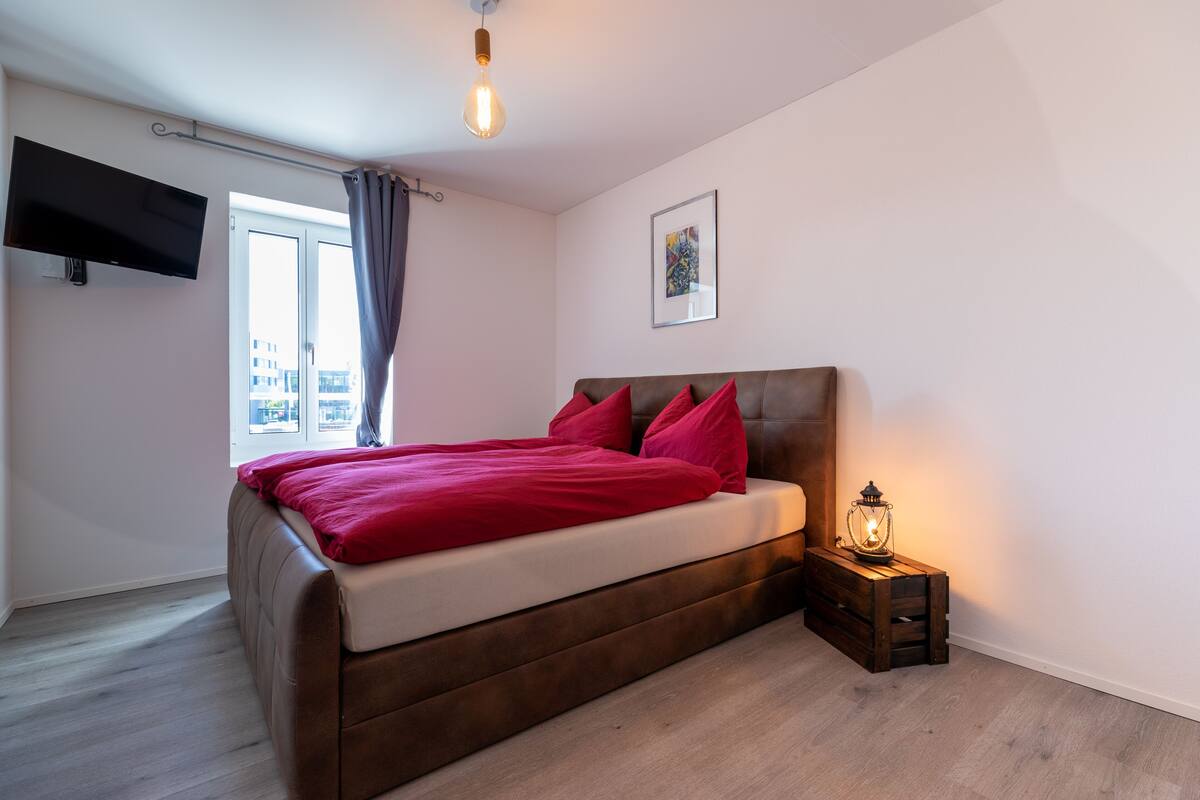 A bedroom with a bed, a TV mounted on the wall, a window with curtains, a bedside table with a lamp, and a framed picture on the wall.