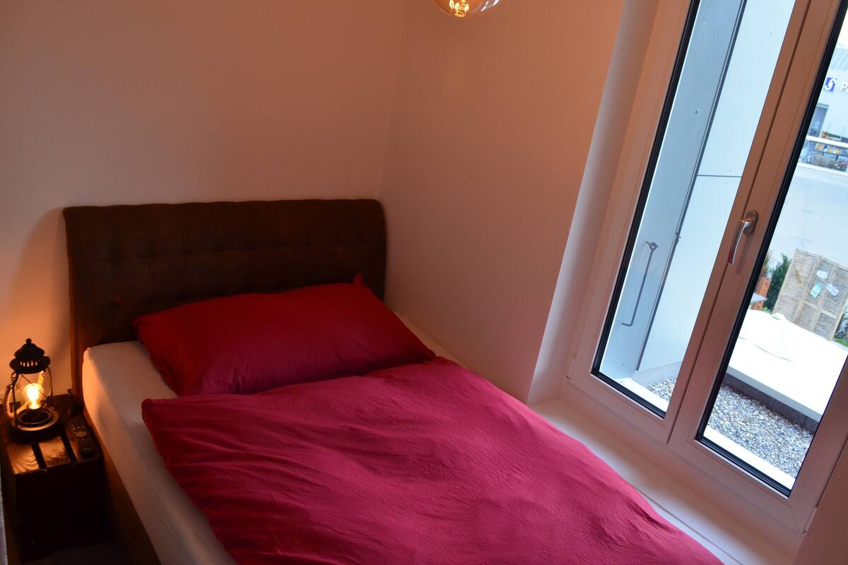 A bed with a red blanket and a headboard, a nightstand with a lamp, and a window with a view of the street.