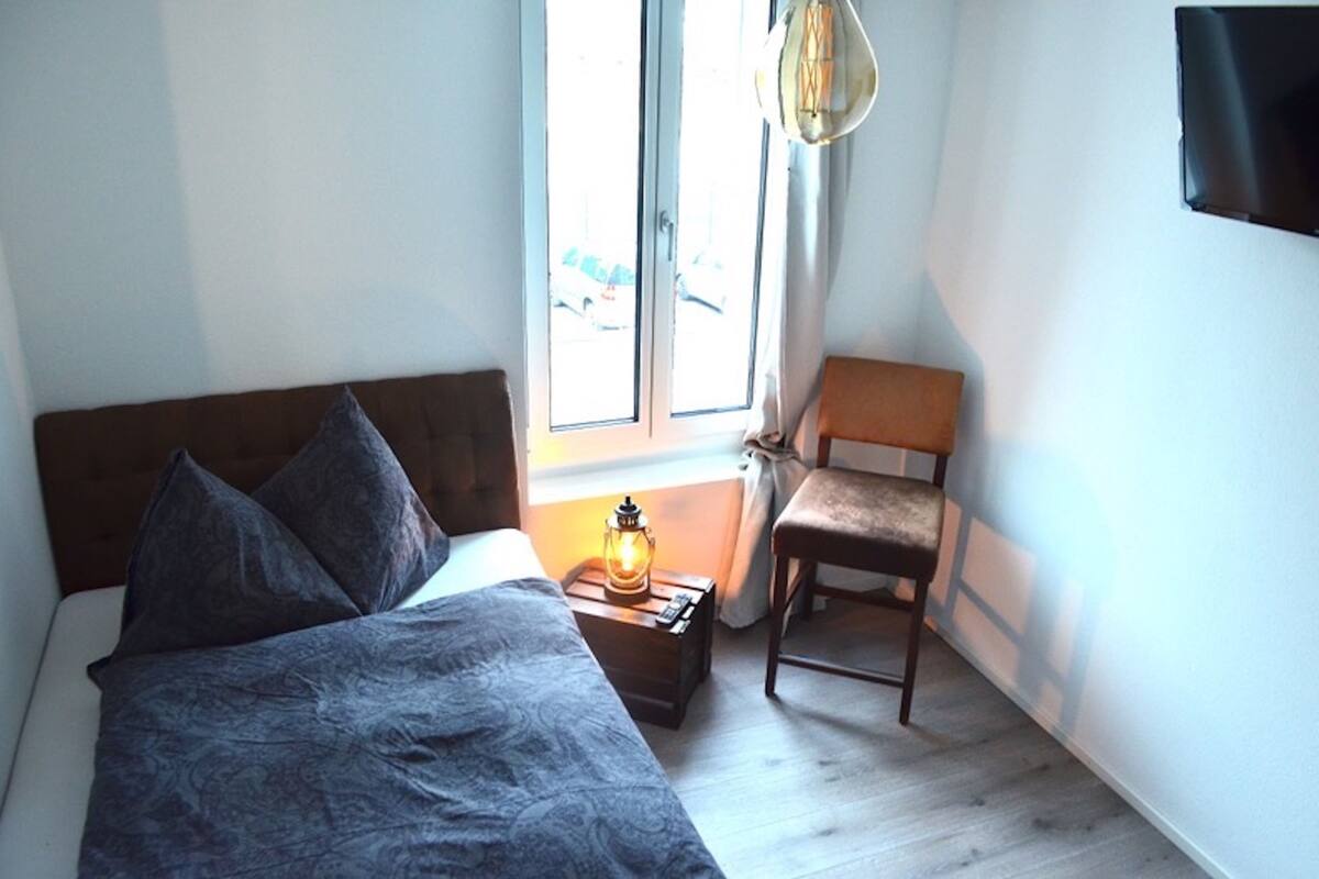 A bedroom with a bed, a nightstand with a lamp, a chair, and a window with curtains.