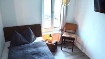 Un dormitorio con cama, una mesita de noche con lámpara, una silla y una ventana con cortinas.