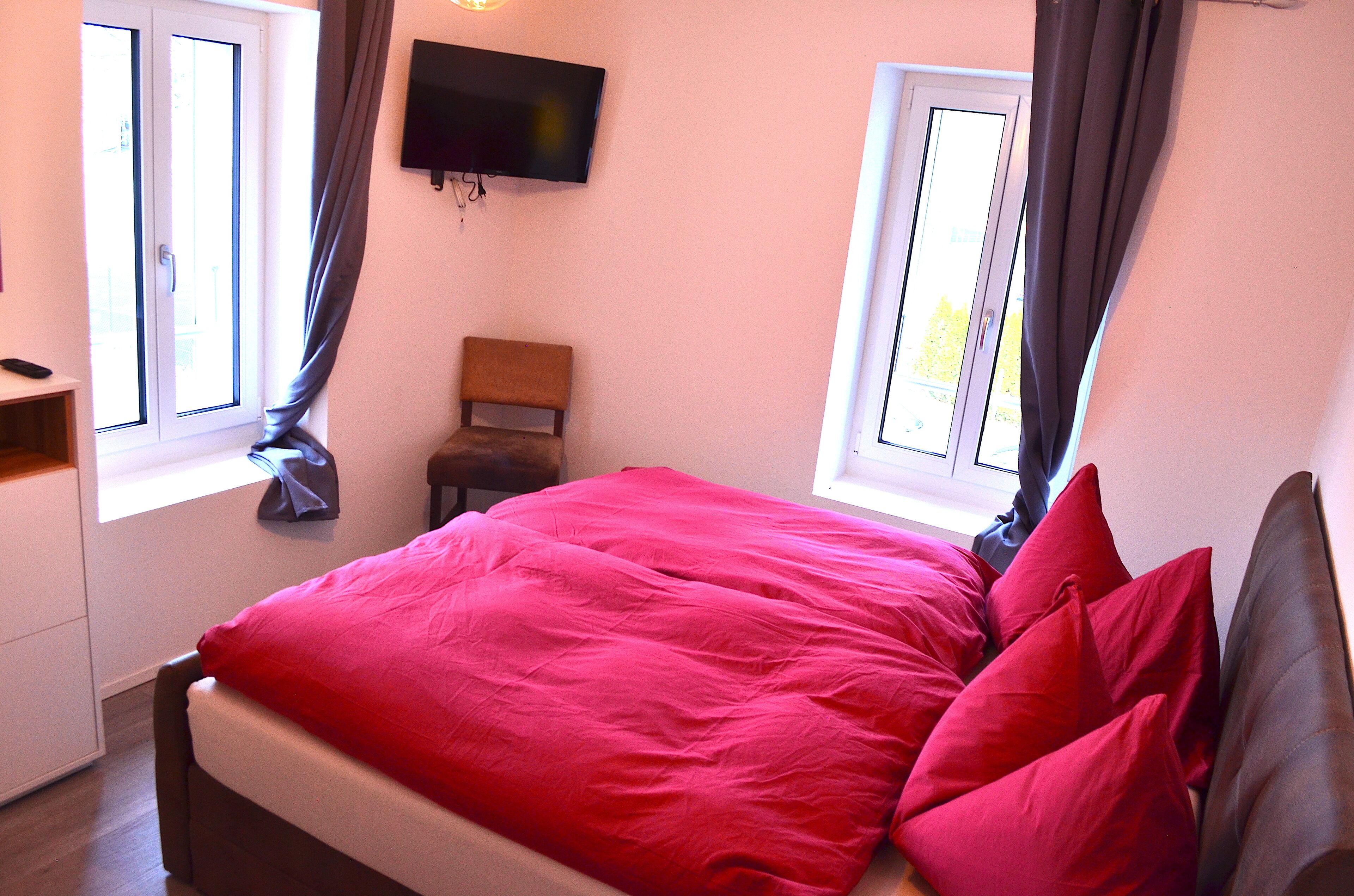 A bedroom with a bed, a chair, a TV, and two windows with curtains.