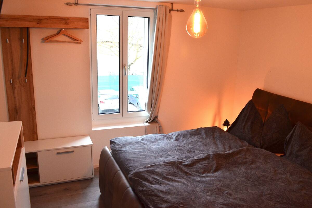 A bedroom with a bed, a window, a hanging light fixture, and a wooden wardrobe.