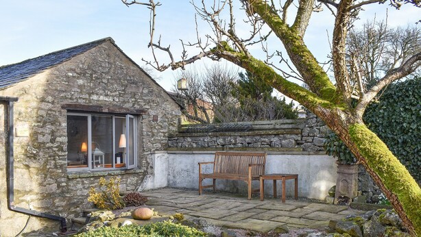 A stone building with a window, a bench, and a small garden area with a hedge and rocks.