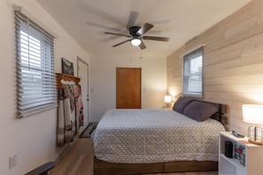 Un dormitorio con cama, ventilador de techo, mesita de noche con lámpara y puerta de madera.