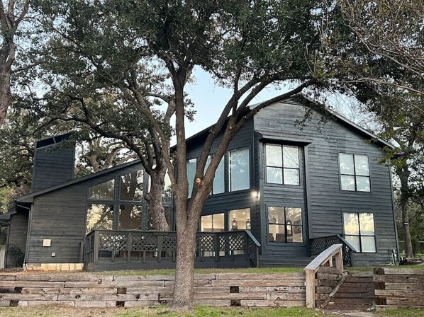 A two-story house with a dark exterior, large windows, and a staircase leading to the entrance. - Huge Lakefront Getaway Hot Tub, Deck, Sleeps 16 (Little Elm)