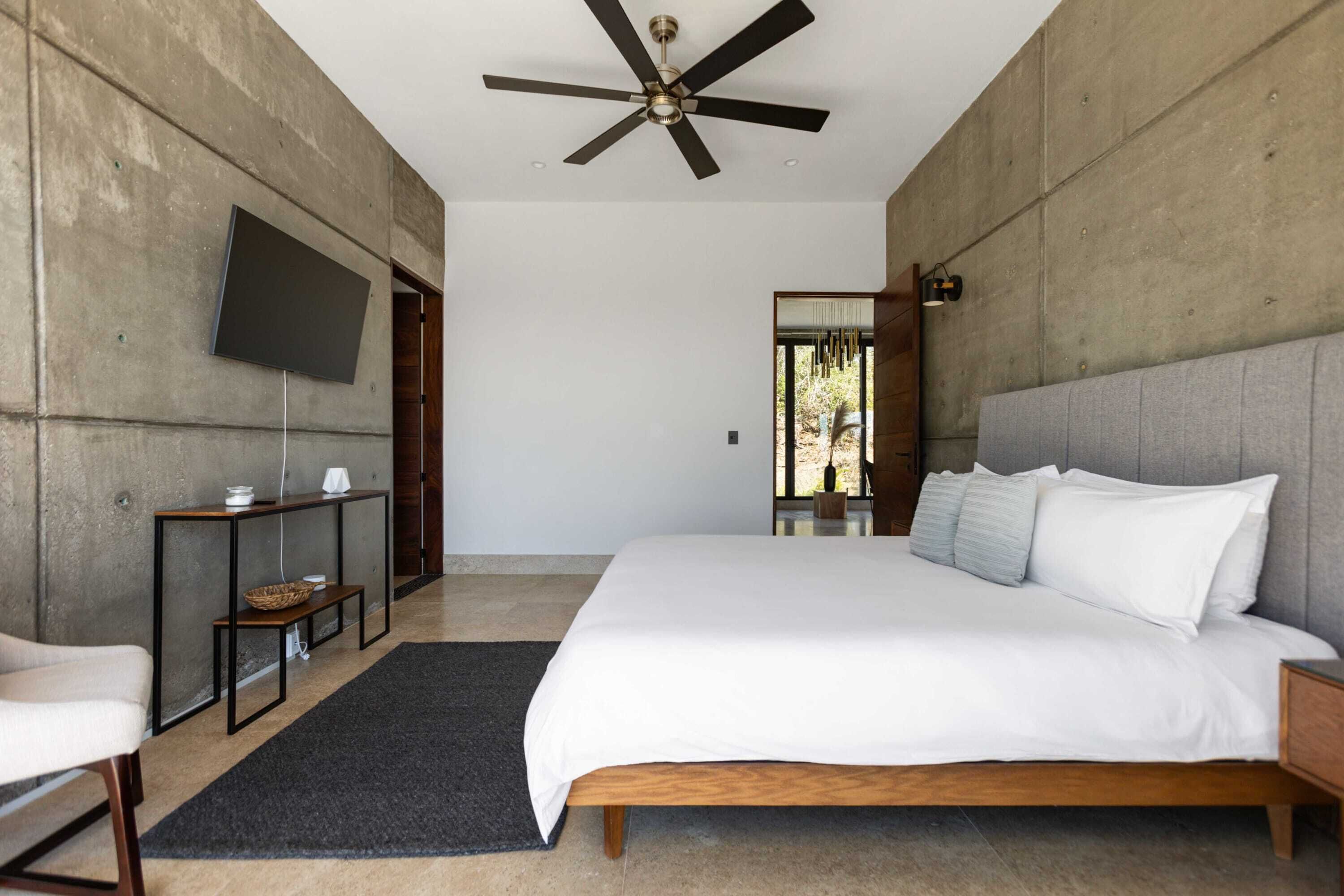 A modern bedroom with a large bed, a ceiling fan, a flat-screen TV mounted on the wall, and a small console table with a basket.