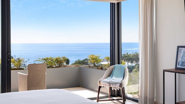 Un balcon avec vue sur l’océan, une chaise en osier et un lit avec une tête de lit blanche.