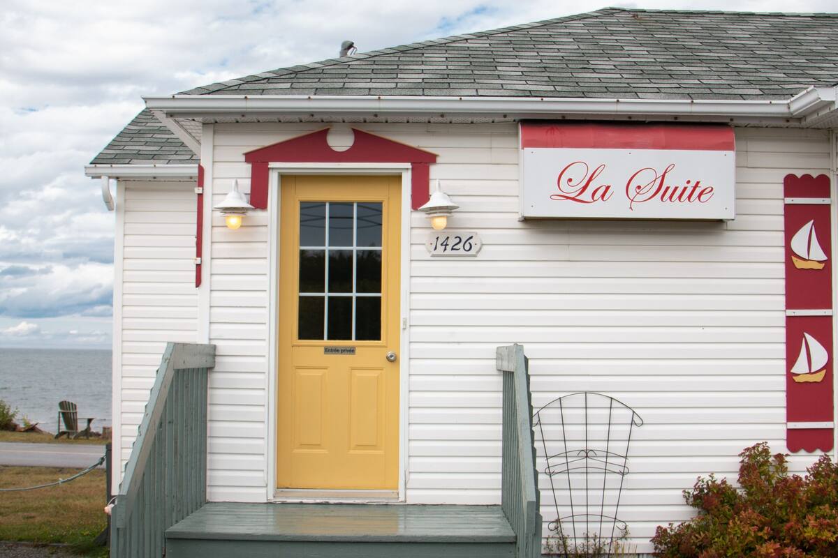 Un bâtiment blanc avec une porte jaune, un auvent rouge et une enseigne sur laquelle on peut lire « La Suite ».