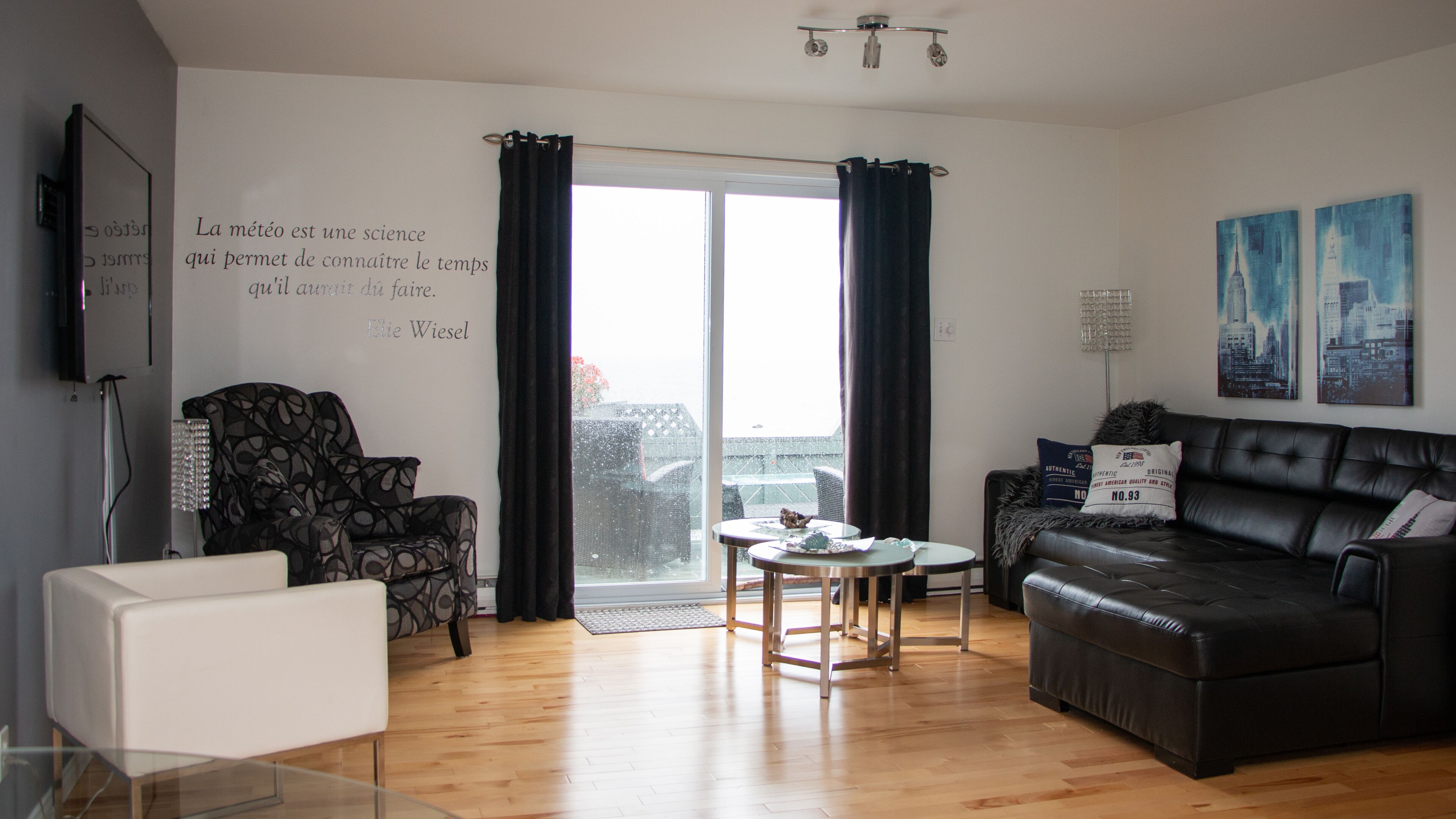 Een moderne woonkamer met een zwarte leren bank, een fauteuil met een patroon en een witte fauteuil. Er is een glazen salontafel en een klein rond tafeltje met een decoratief object. De kamer heeft houten vloeren en grote ramen met donkere gordijnen.